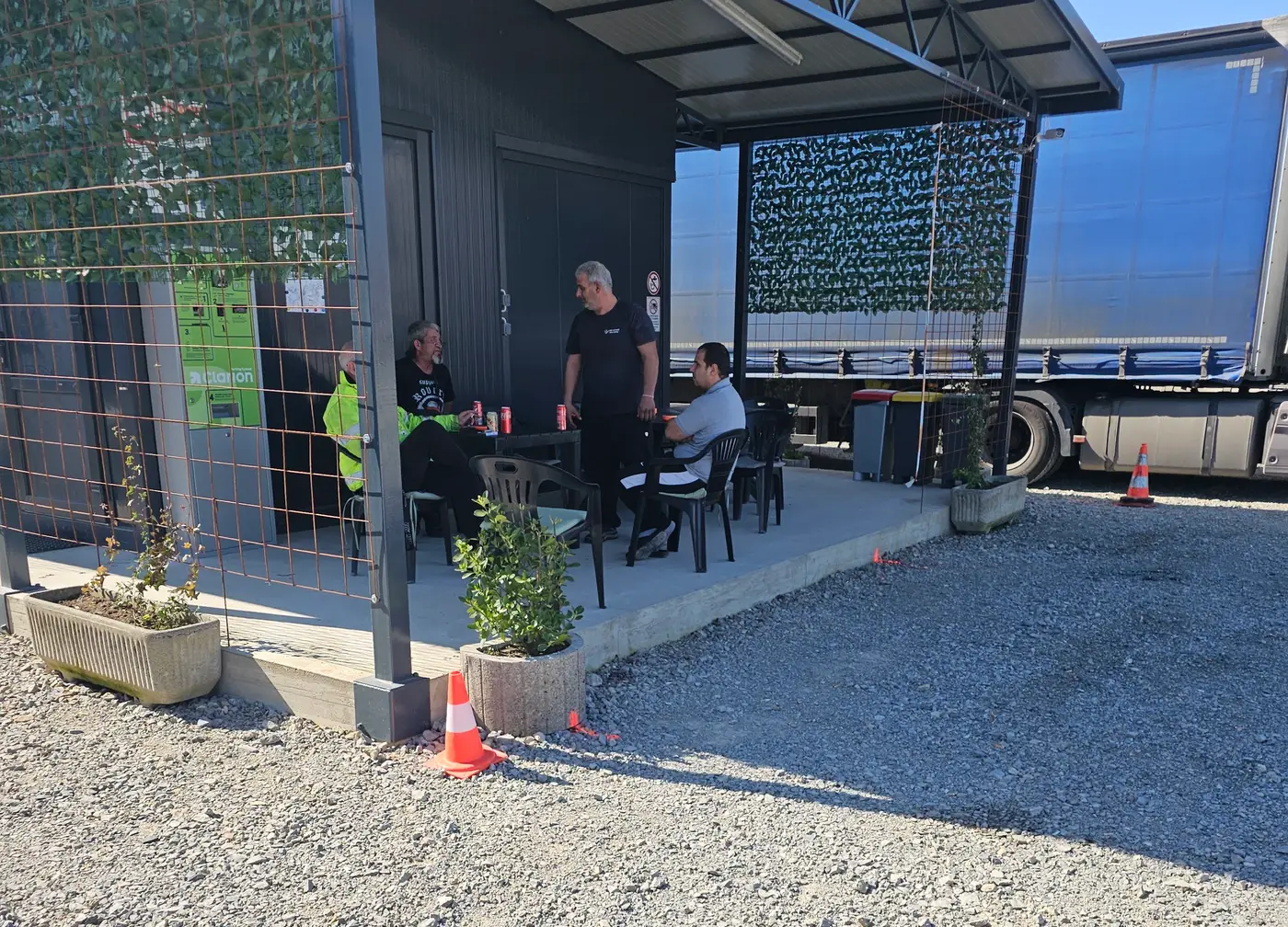 Vozači sjede za stolom na natkrivenoj terasi parkinga, iza njih parkirana poluprikolica
